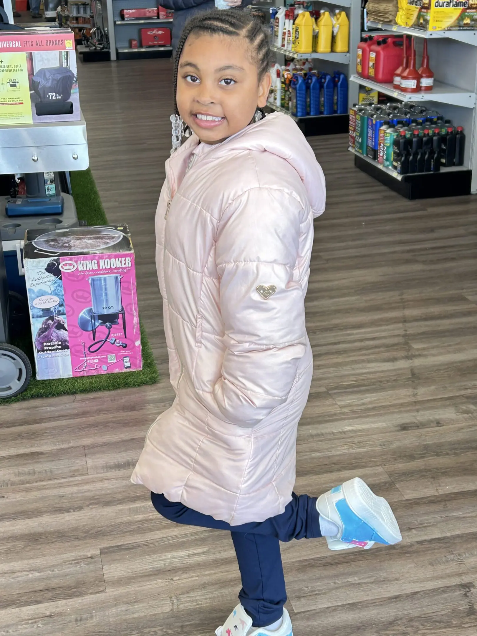 A child in a pink coat and blue jeans stands indoors with a playful smile.